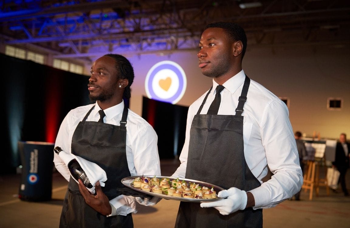 Two servers, one holding a bottle of champagne while the other serves canapés